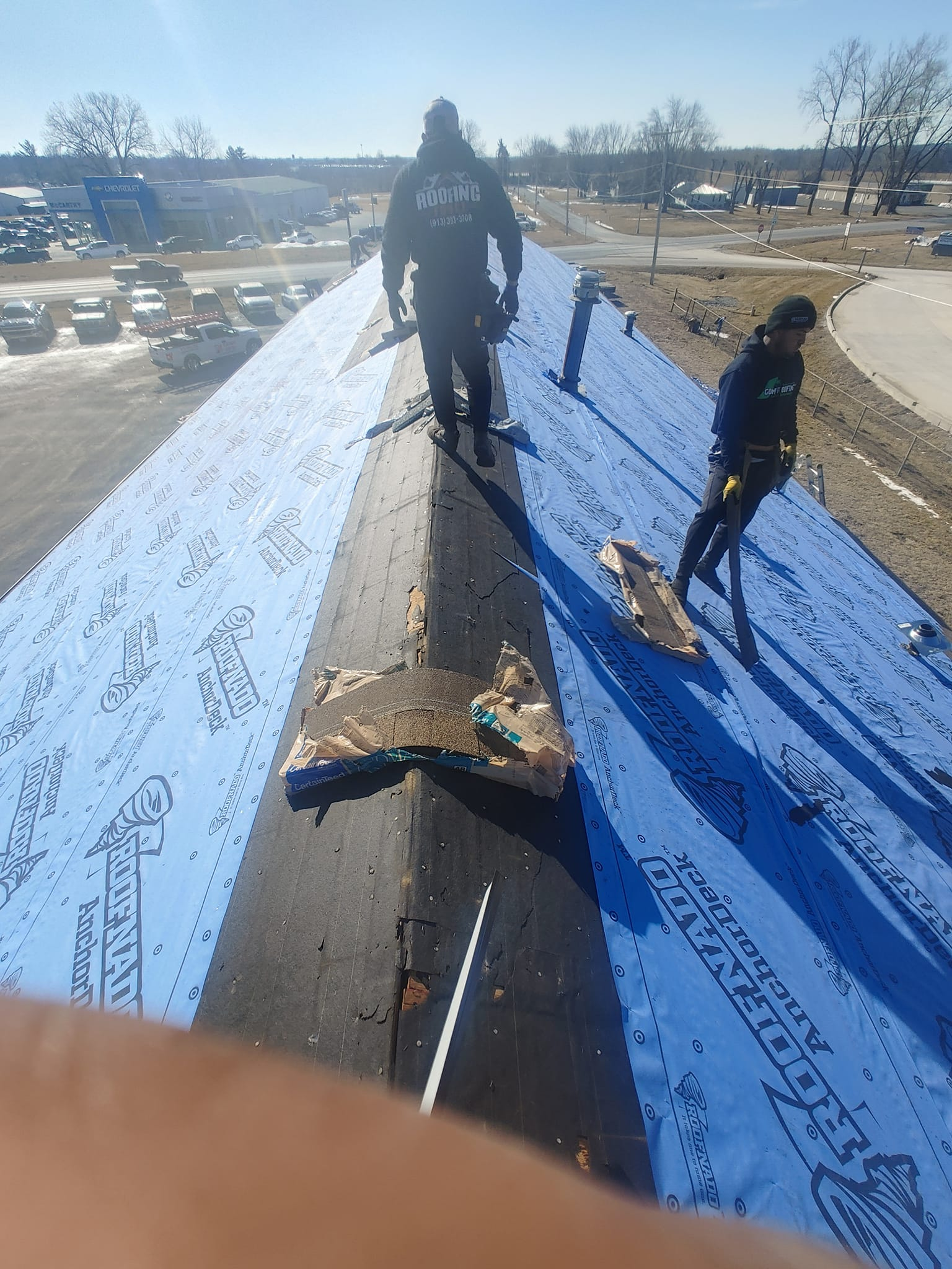 Roof replacement in progress with two workers on a sloped commercial roof, new shingles stacked on roof pitch, chevrolet car dealership in background